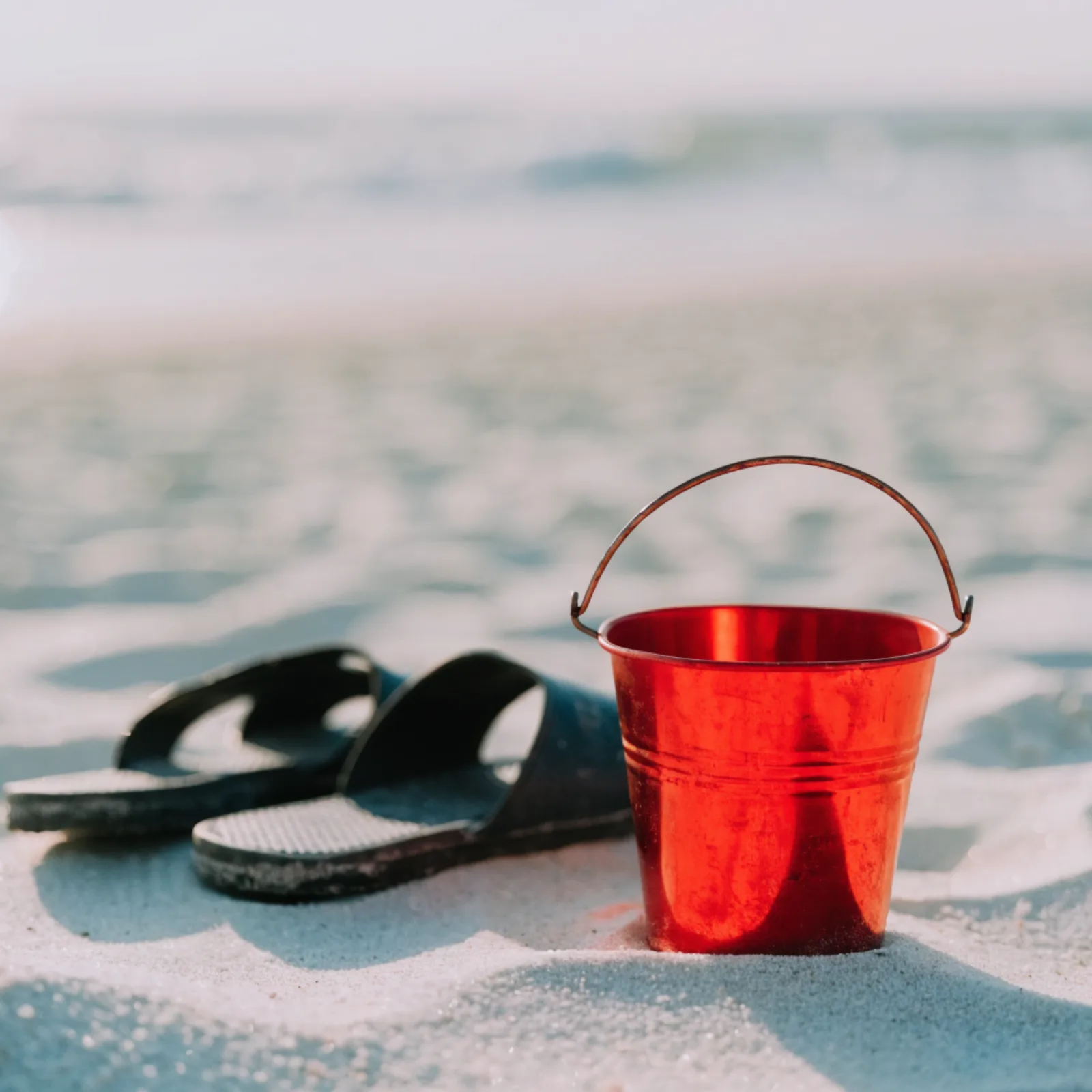 A small red tin bucket and sandals on powder-white sand