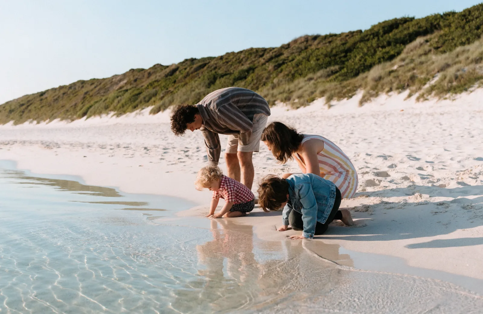 Parents and kids playing at the water's edge below a tall dune ridge